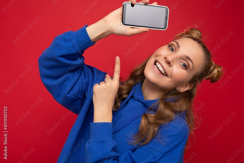 Naklejka premium Attractive smiling young blonde woman wearing stylish blue hoodie isolated on red background with copy space holding smartphone showing phone in hand with empty screen display pointing at gadjet