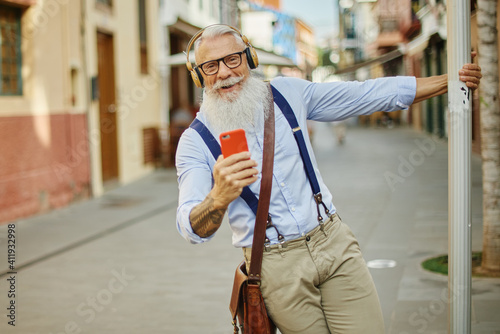 Older hipster man using a smartphone and playing music, having fun with mobile phone technology while holding on to a traffic sign in the city - Happy, technological and cheerful senior lifestyle 
