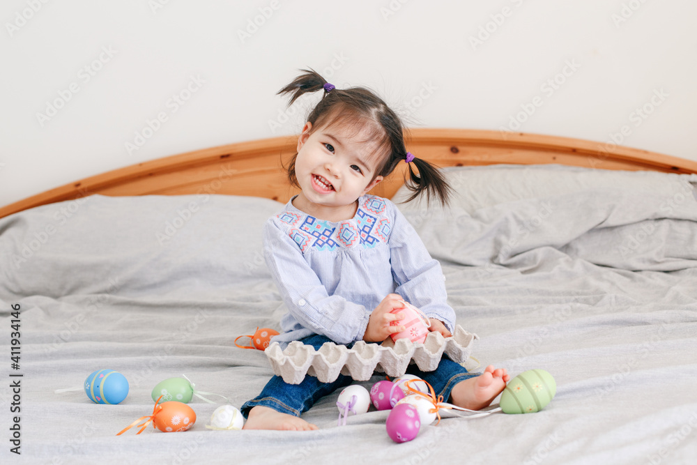 Obraz premium Happy Easter. Asian Chinese baby girl with funny pigtails sitting on bed at home and playing with colorful Easter eggs. Kid child celebrating a traditional Christian holiday.