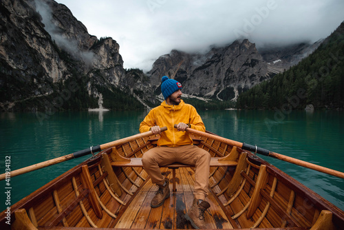Traveler man with blue hat and yellow raincoat sailing in a wooden boat through a beautiful Di Braies lake in the Dolomites mountains. 