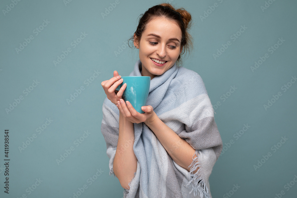 Shot of beautiful young cute smiling brunette woman wearing warm blue scarf isolated over blue background holding blue mug drinking coffee and looking down Shot of beautiful young cute smiling brunette woman wearing warm blue scarf isolated over blue background holding blue mug drinking coffee and looking down
