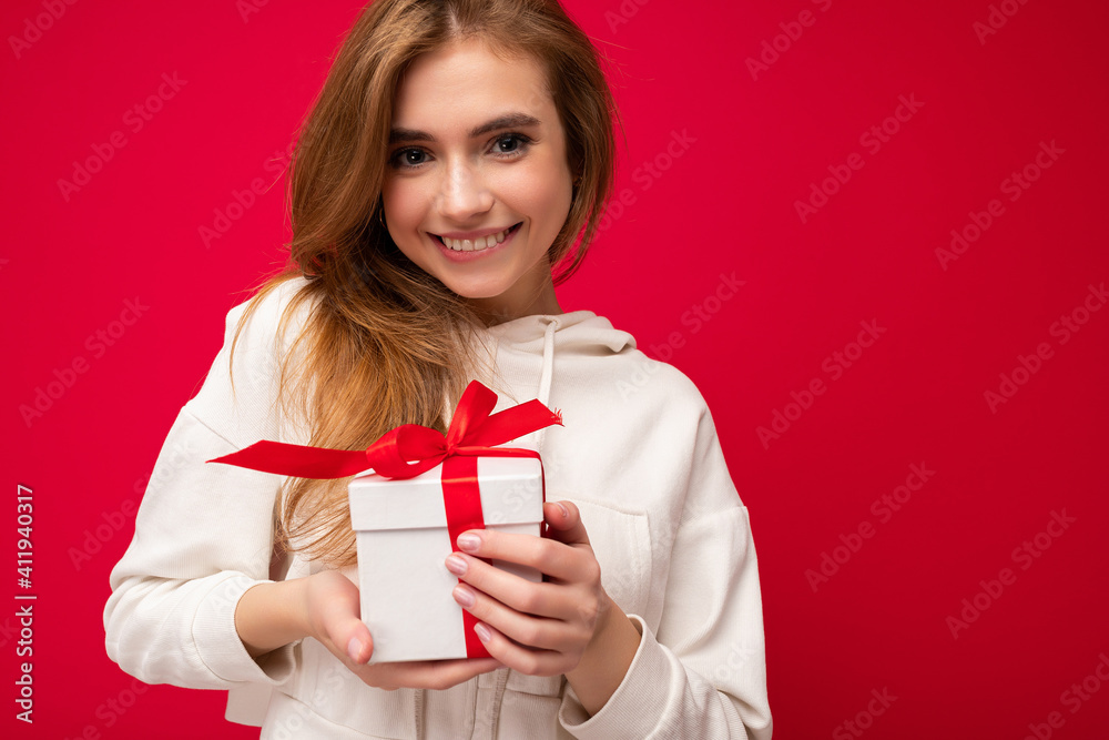 Photo shot of attractive happy smiling cute blonde young female person isolated over red background wall wearing white hoodie holding white gift box with red ribbon and looking at camera Photo shot of attractive happy smiling cute blonde young female person isolated over red background wall wearing white hoodie holding white gift box with red ribbon and looking at camera