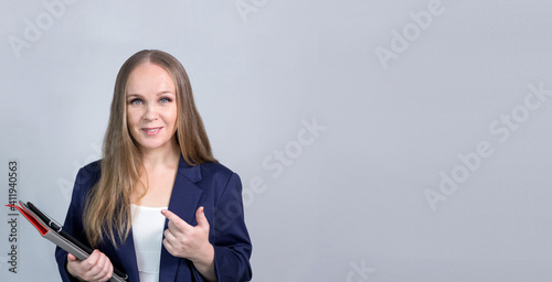 Banner. A business girl on a light background with a tablet computer stands, smiles, wants to say. She is wearing a blue jacket and a white blouse, with long brown hair.