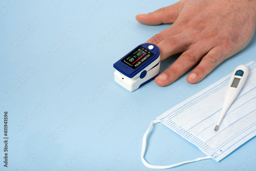 Human male hands pulse oximeter used to measure pulse rate and oxygen levels with medical blue background with copy space. Top view. Medical concept protective medical mask, electronic thermometer. Human male hands pulse oximeter used to measure pulse rate and oxygen levels with medical blue background with copy space. Top view. Medical concept protective medical mask, electronic thermometer.