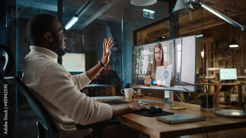 Handsome Black African American Project Manager is Making a Video Call on Desktop Computer in a Creative Office Environment. Male Specialist Talking to a Caucasian Female Colleague Over a Live Camera.