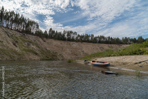 Stop for a kayak on the low bank. It's raining. The river makes a turn. The kayak has a backpack and an aladka. Pine trees on the steep right bank. Summer tourist parking. Solo hike.