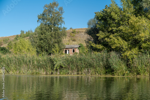 An abandoned house with gaping windows on the banks of the Khoper River. On the edge of the coast there is reed, birch. Hill. photo