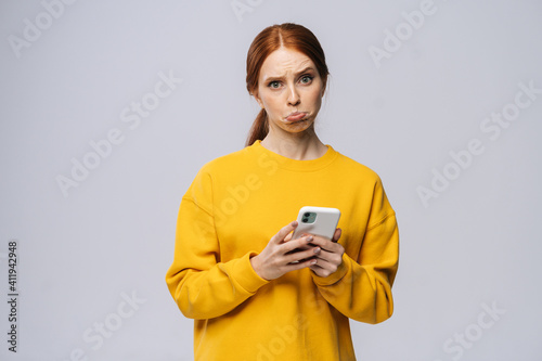 Displeased cute young woman reading bad news, feels dissatisfaction after receiving message and looking at camera on isolated white background. Pretty lady with red hair showing facial expressions © dikushin