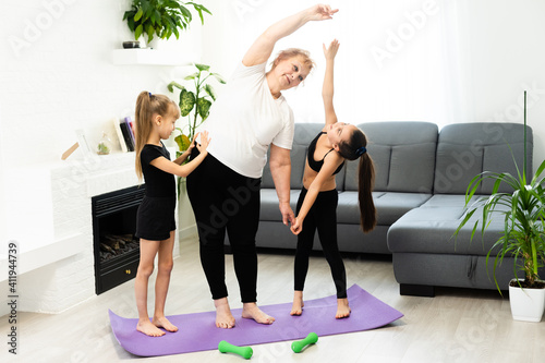 senior grandmother workout yoga withgranddaughters preschooler. They are sitting on mat at cozy home interior. Sport, parenthood and people concept