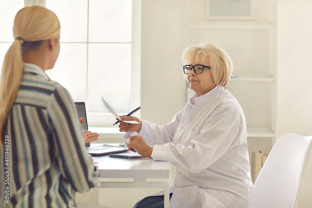 Obraz premium Friendly senior female doctor gives a patient a prescription while sitting at a table in the clinic office. Young woman talking to family doctor sitting with her back to the camera. Healthcare concept