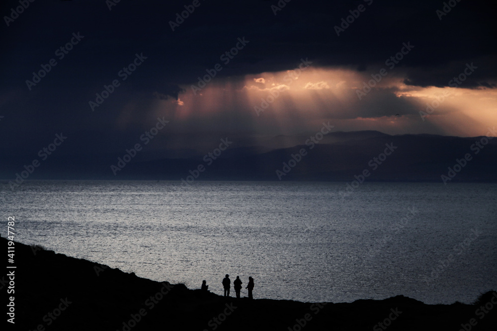 Four people backlit in an oceanfront sunset scene. Sunbeams break through the clouds at sunset. Cabo de Gata Park. Almeria - obrazy, fototapety, plakaty 