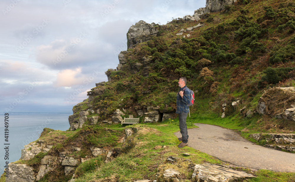 A hiker with a backpack stops on the coastal path and looks out to sea. The trail is in Somerset in the south of England. In the background are the Bristol Channel and a sky with clouds.