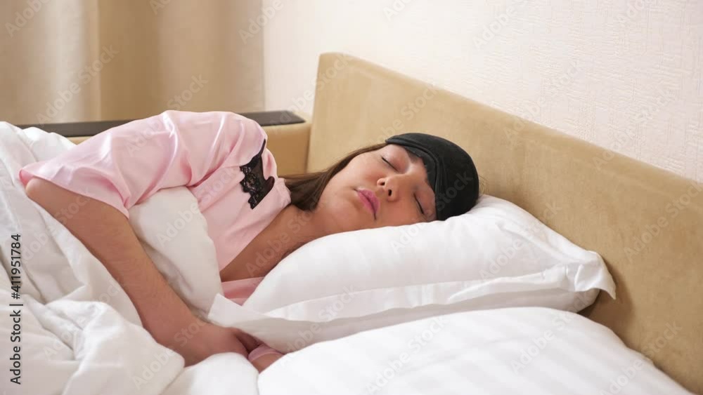 Young woman wearing sleeping eye mask lies relaxed in modern bed with light linen in bedroom closeup Young woman wearing sleeping eye mask lies relaxed in modern bed with light linen in bedroom closeup