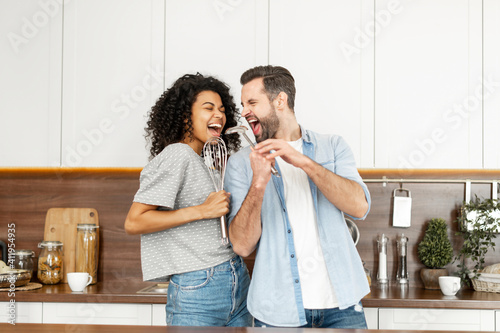 Obraz na plátně Happy interracial couple dancing in the kitchen, singing while cooking breakfast