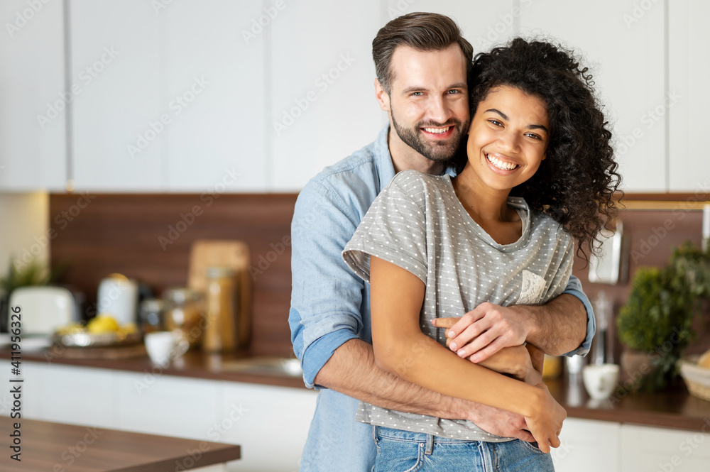 Smiling man hugging from behind charming African American woman, two people standing and joyfully looking at camera. Young international couple happily spending time in cozy modern kitchen at home. Smiling man hugging from behind charming African American woman, two people standing and joyfully looking at camera. Young international couple happily spending time in cozy modern kitchen at home.