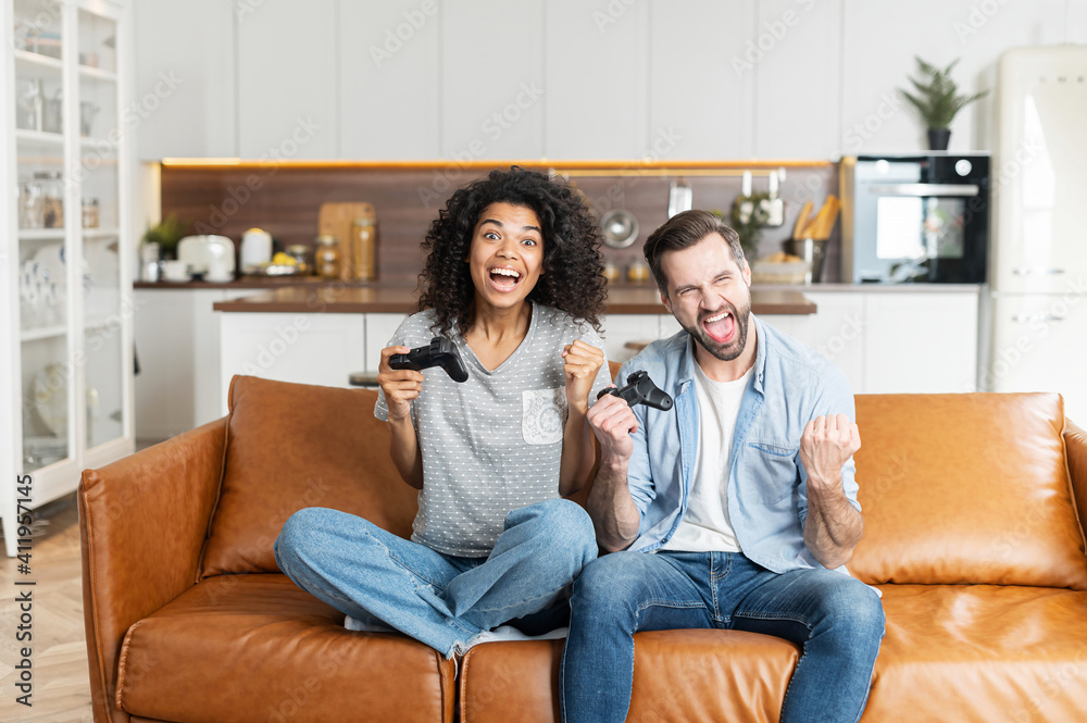 Young African American woman and handsome man sitting on the couch, interracial couple boyfriend and girlfriend playing video game with joysticks in living room at home, winning a game and gesturing Young African American woman and handsome man sitting on the couch, interracial couple boyfriend and girlfriend playing video game with joysticks in living room at home, winning a game and gesturing
