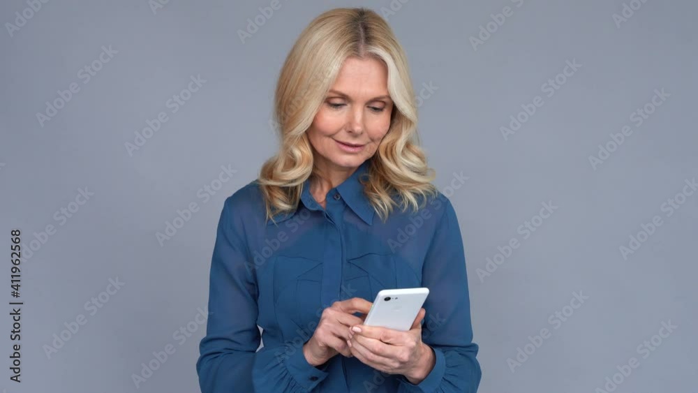 Happy middle aged business woman using apps looking at smartphone standing on grey wall. Smiling 50s older lady holding phone advertising online mobile e services, texting, searching applications.