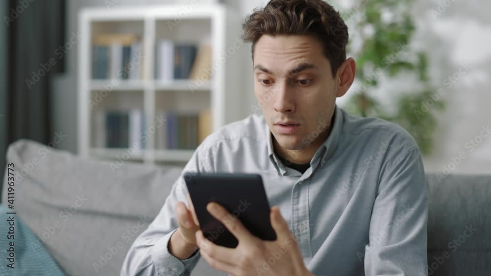 Surprised man in casual shirt looking at tablet screen while sitting on grey couch. Young guy spending free time at home with modern device. Surprised man in casual shirt looking at tablet screen while sitting on grey couch. Young guy spending free time at home with modern device.