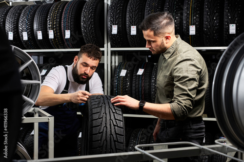 auto mechanic talk about advantages of auto tire to young client in service, man came to buy new tire for his automobile, stand talking and examining the product