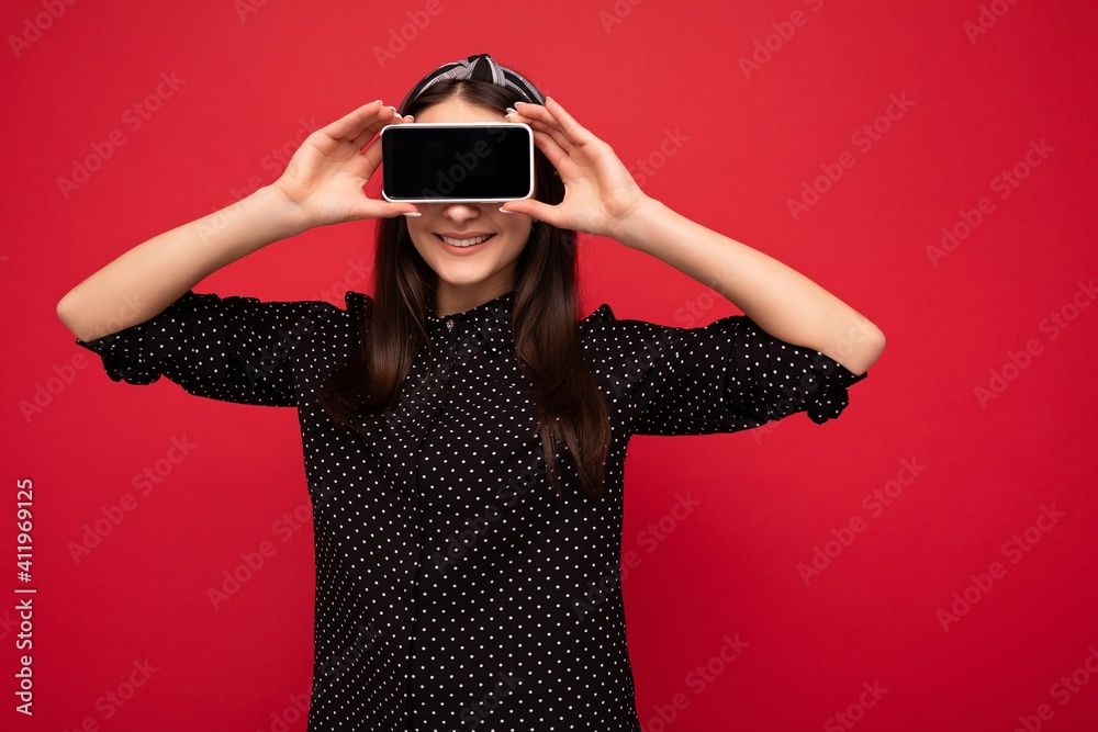 Pretty smiling joyful brunette girl standing isolated over red wall wearing casual stylish black clothes showing mobile phone with empty screen for cutout Pretty smiling joyful brunette girl standing isolated over red wall wearing casual stylish black clothes showing mobile phone with empty screen for cutout