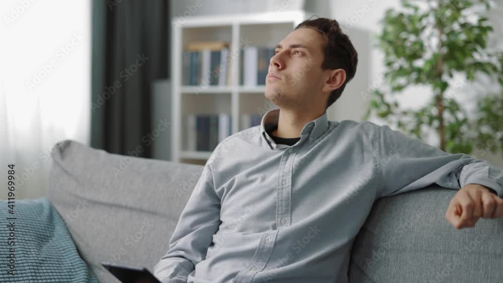 Thoughtful man sitting on couch, holding digital tablet and looking aside. Young guy in casual shirt spending free time online while staying at home. Thoughtful man sitting on couch, holding digital tablet and looking aside. Young guy in casual shirt spending free time online while staying at home.