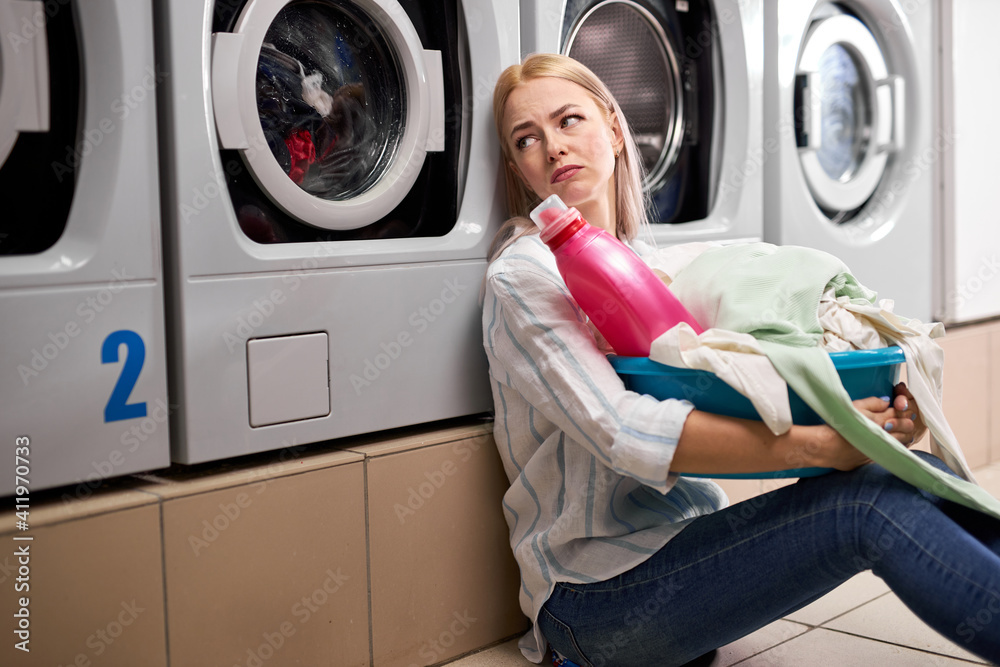 Bored woman holding dirty clothes and detergent pink bottle in basin, sits on the floor leaning on washing machine, sit depressed alone, in washing house Bored woman holding dirty clothes and detergent pink bottle in basin, sits on the floor leaning on washing machine, sit depressed alone, in washing house