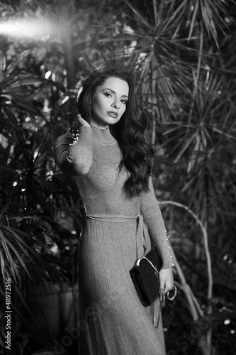 Long-haired woman in gray knitted dress standing against tropical trees on background