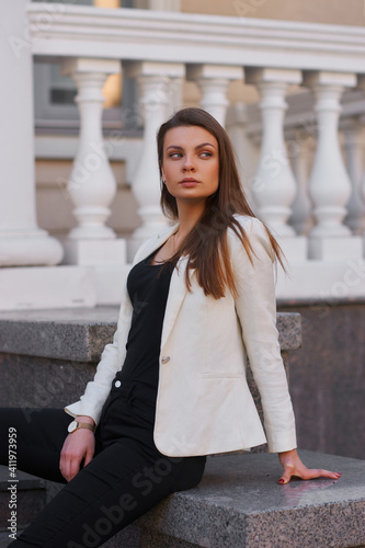 Young woman in black shirt, jeans and white blazer walking and posing outdoors on a summer day. Sun backlight. Outdoor portrait