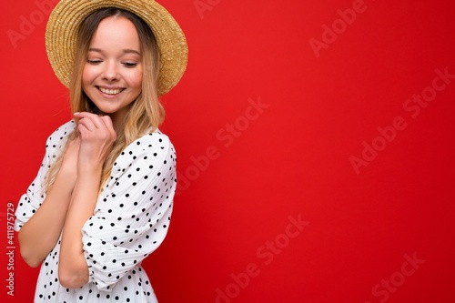 Portrait of young beautiful smiling hipster blonde woman in trendy summer dress and straw hat. Sexy carefree female person posing isolated near red wall in studio. Positive model with natural makeup