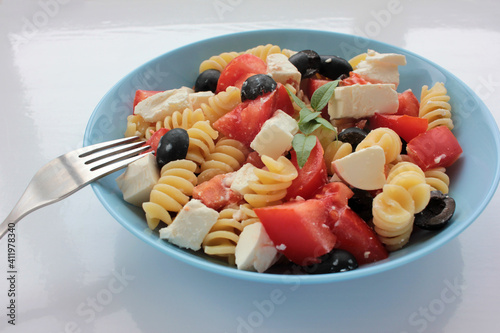 Itlian fusilli pasta salad with olives, tomato, and  feta cheese in blue bowl on white table background. Overhead view photo