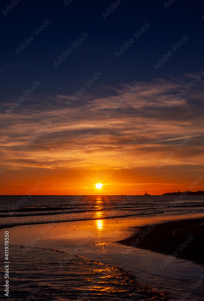 The beautiful sunset, orange blue sky and clouds over the Blyth Beach. St. Mary's Lighthouse and Seaton Sluice village  in the background. Cold winter day in Blyth, Northumberland, UK. Vertical image.