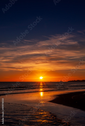 The beautiful sunset, orange blue sky and clouds over the Blyth Beach. St. Mary's Lighthouse and Seaton Sluice village in the background. Cold winter day in Blyth, Northumberland, UK. Vertical image.
