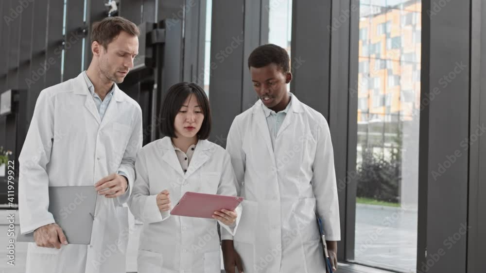 Medium shot of two diverse male doctors and their female Asian colleague going along modern hospital and discussing information from tablet woman holding in hands Medium shot of two diverse male doctors and their female Asian colleague going along modern hospital and discussing information from tablet woman holding in hands