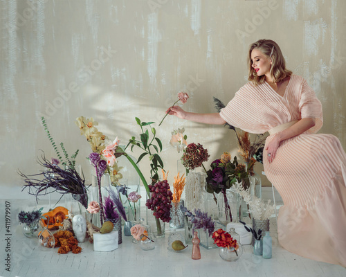an elegant blonde girl in a beautiful light pink dress sits surrounded by gorgeous flowers on a light background