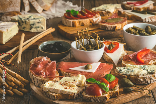 Baguette bread slices with cheese, bacon, salami, olives and capers as a starter, tapas, antipasti, mediterranean style on a rustic wooden table
