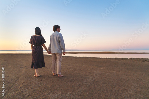 Young couple of lovers in linen clothes walking along the evening beach. Copy space.