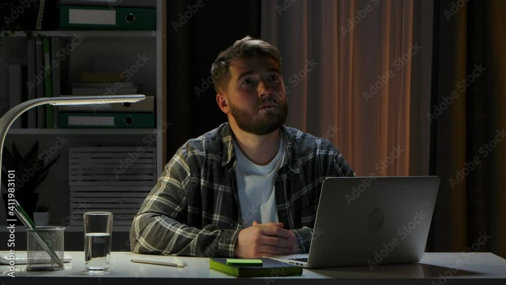 Tired young man sits at a laptop in a dark home office and sighs heavily, upset with no more ideas. Remote work from home. End of the working day. Freelance. Online training. Close up. Slow motion. Tired young man sits at a laptop in a dark home office and sighs heavily, upset with no more ideas. Remote work from home. End of the working day. Freelance. Online training. Close up. Slow motion.