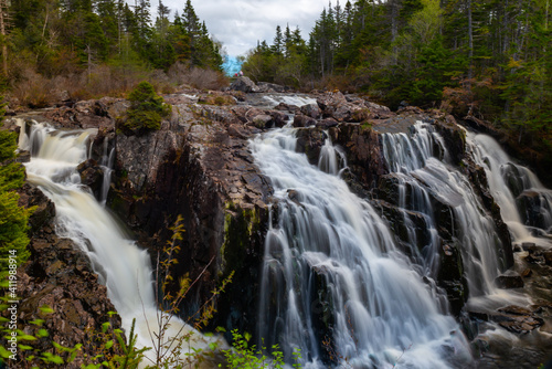 A raging river of white rapids and waterfalls with tall evergreen trees on both sides. The stream is enclosed by large boulders or rock formations with dead read leaves and moss covering greenery. 