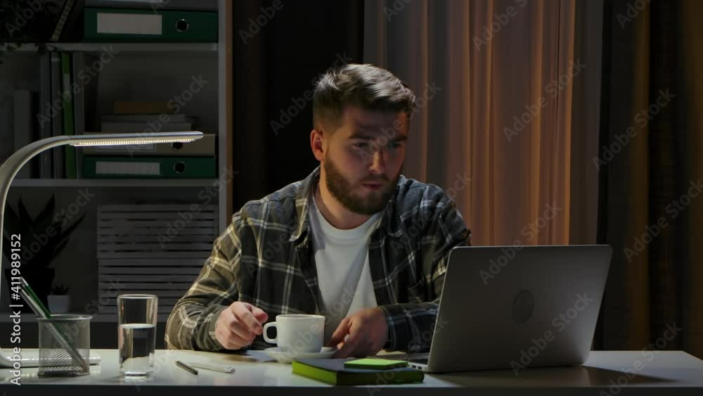 Creative man sitting at his desk in home office studio, working on laptop in the evening. The remote worker works and enjoys hot coffee. Remote work, online training at home. Close up. Slow motion. Creative man sitting at his desk in home office studio, working on laptop in the evening. The remote worker works and enjoys hot coffee. Remote work, online training at home. Close up. Slow motion.