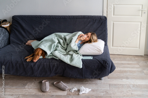 Sick woman at home lying in bed with her Vizsla dog, suffering from allergy, rhinitis or flu symptom, covering with plaid and blowing her nose, sneeze in paper tissue. Cold and seasonal illnesses. 