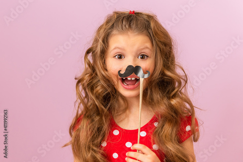 A little girl with curly hair holds a fancy paper moustache.