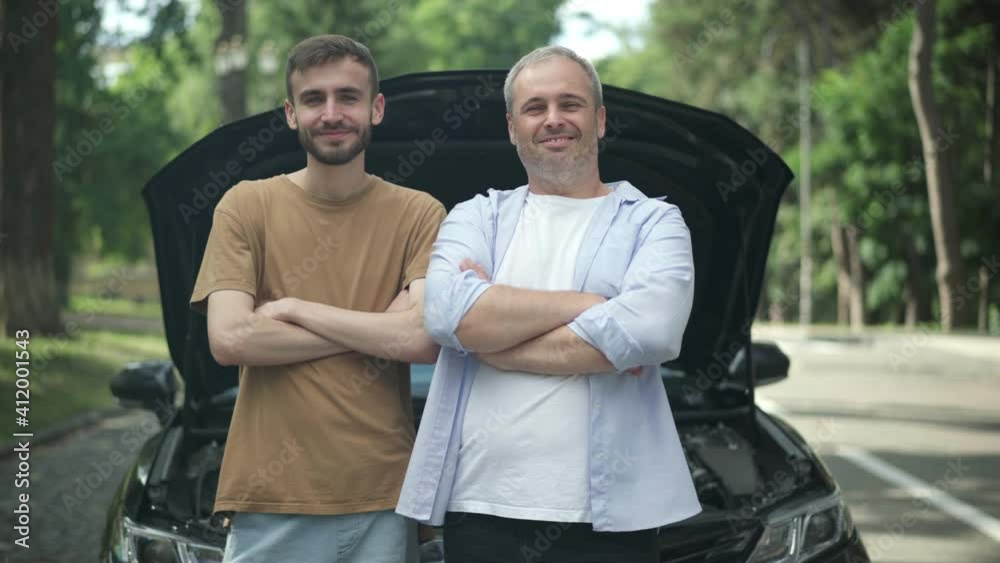 Confident smiling middle aged and young men standing at open car hood crossing hands and looking at camera. Portrait of joyful Caucasian father and son posing with broken automobile on summer road. Confident smiling middle aged and young men standing at open car hood crossing hands and looking at camera. Portrait of joyful Caucasian father and son posing with broken automobile on summer road.