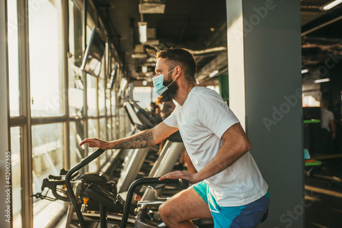 An active man in a protective medical mask training in gym, riding a stationary exercise bike. Sport and quarantine.