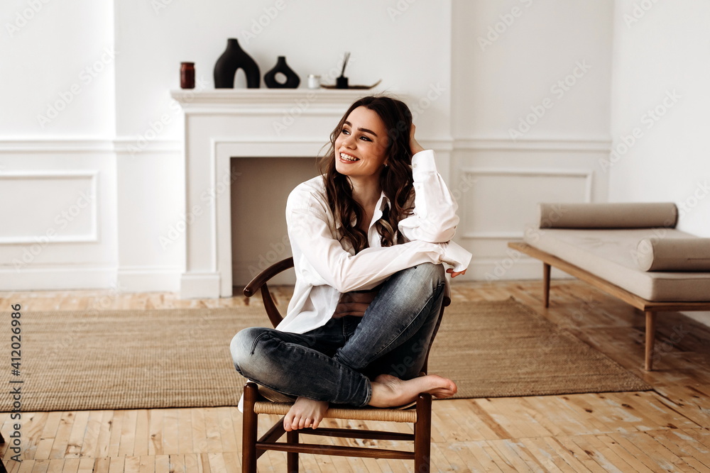 happy woman of European appearance in a white shirt smiling. Brunette lady laughs while sitting on a chair in a beautiful interior, close-up portrait. 