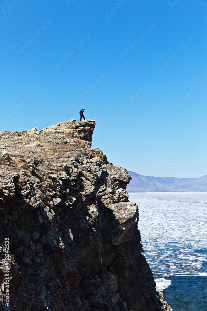Baikal Lake in sunny May day. A tourist photographs an ice drift in the Small Sea Strait from a high cliff. Spring travel and active holidays