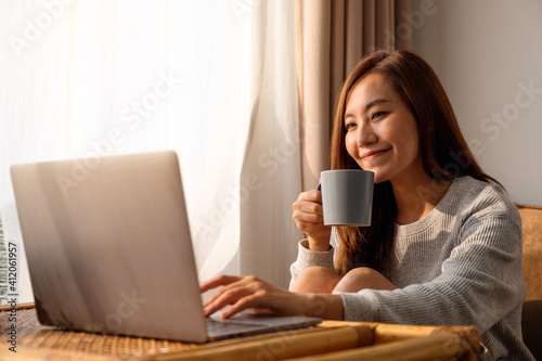 A beautiful young asian woman using and working on laptop computer while drinking coffee at home