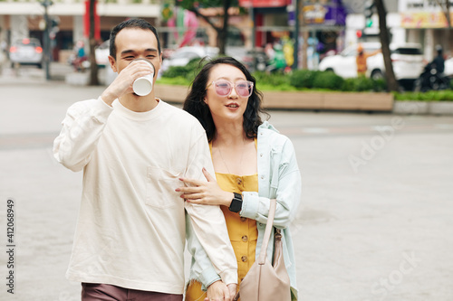 Happy Chinese young man drinking take out coffee when walking with beautiful girlfriend on the street