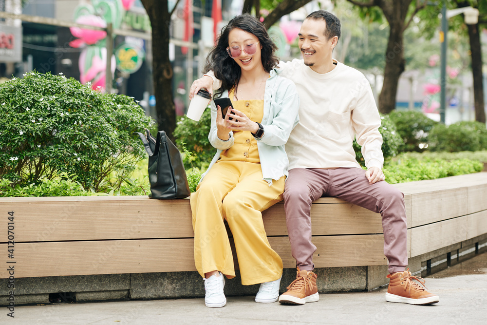Happy young Chinese couple sitting on bench and checking social media on smartphone or posting photos
