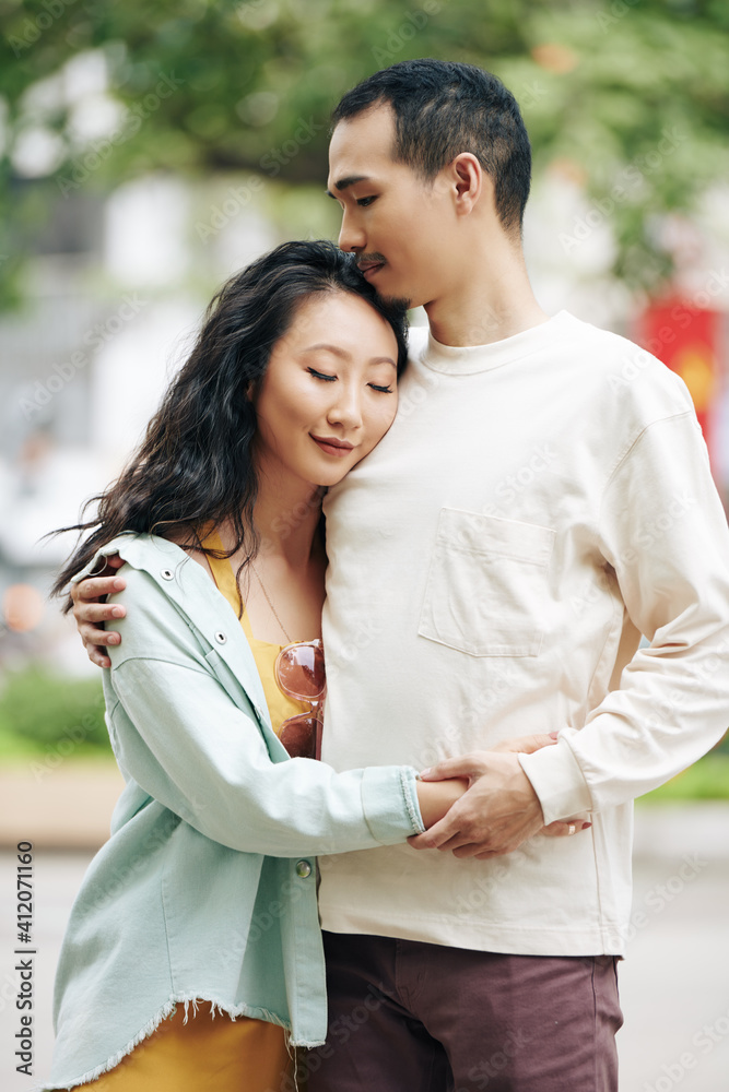 Happy young Chinese couple in love in casual clothes standing outdoors, hugging and closing eyes Happy young Chinese couple in love in casual clothes standing outdoors, hugging and closing eyes