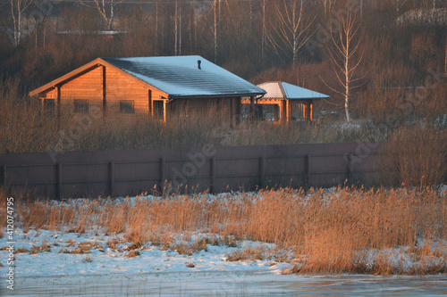 Winter rural landscape at dawn of a cold day. A new wooden cattage on the banks of a frozen river or lake among trees and dry reeds. photo